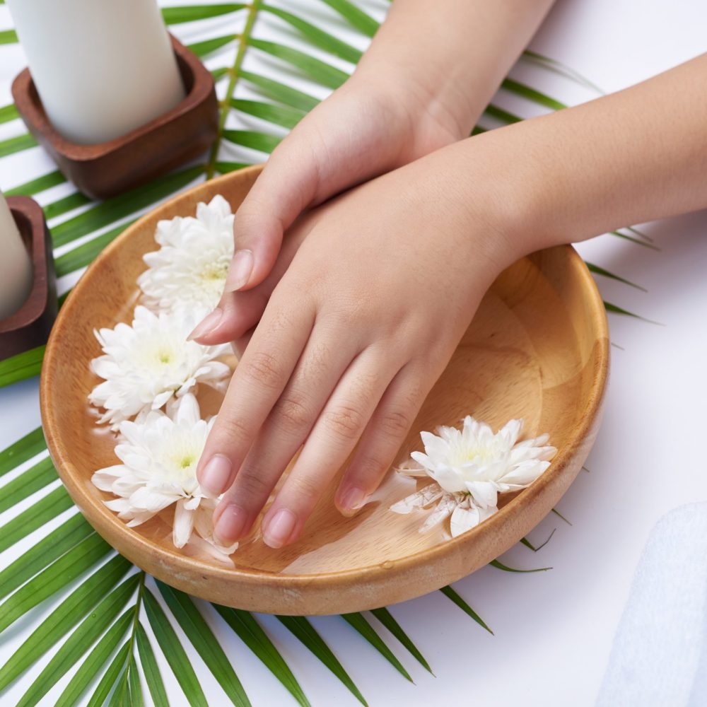Woman soaking her hands in bowl of water and flowers, Spa treatment and product for female feet and hand spa, massage pebble, perfumed flowers water and candles, Relaxation. Flat lay. top view.